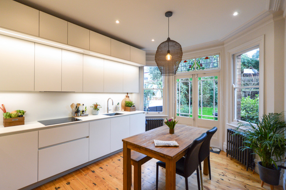 Photo of a mid-sized contemporary l-shaped eat-in kitchen in London with a drop-in sink, flat-panel cabinets, white cabinets, quartzite benchtops, stainless steel appliances, light hardwood floors, no island and white benchtop.