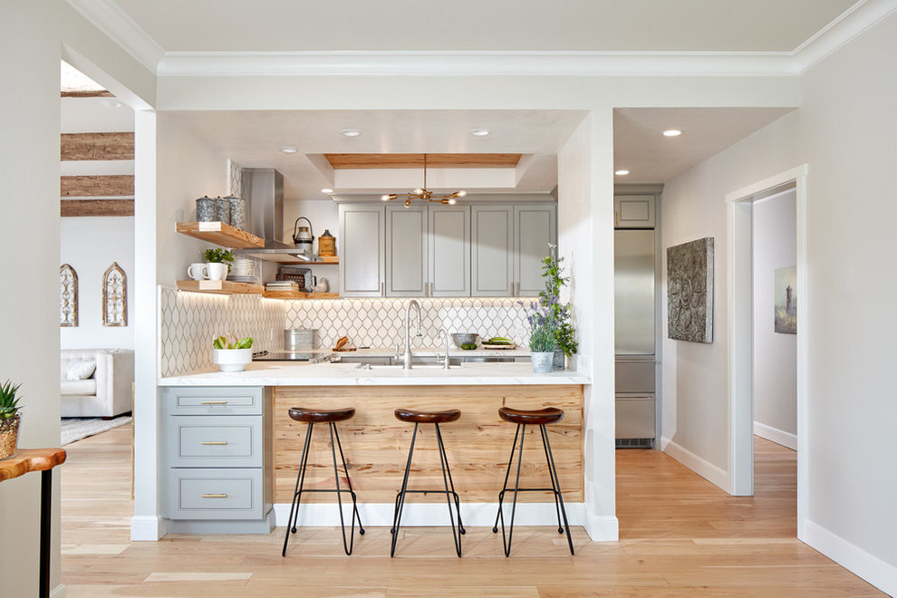 Inspiration for a beach style u-shaped kitchen in San Francisco with recessed-panel cabinets, grey cabinets, white splashback, stainless steel appliances, light hardwood floors, a peninsula, beige floor, white benchtop, a farmhouse sink, marble benchtops and ceramic splashback.