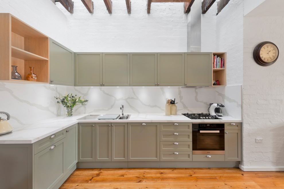 Mid-sized transitional l-shaped eat-in kitchen in Melbourne with a drop-in sink, shaker cabinets, green cabinets, quartz benchtops, white splashback, stone slab splashback, black appliances, medium hardwood floors, with island, brown floor and white benchtop.