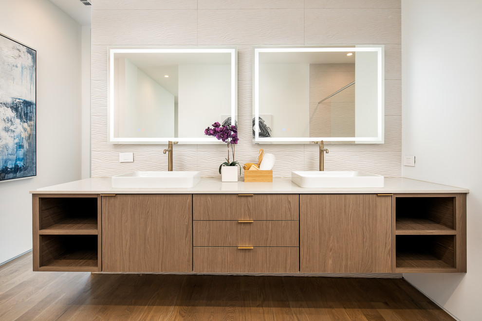 This is an example of a large modern master wet room bathroom in Dallas with flat-panel cabinets, medium wood cabinets, a freestanding tub, a one-piece toilet, beige tile, white walls, medium hardwood floors, a vessel sink, engineered quartz benchtops, brown floor, a hinged shower door and white benchtops.
