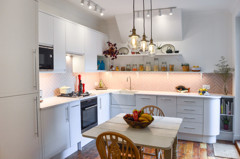 Photo of a contemporary l-shaped eat-in kitchen in London with a farmhouse sink, raised-panel cabinets, white cabinets, pink splashback, panelled appliances, medium hardwood floors, no island, brown floor and white benchtop.