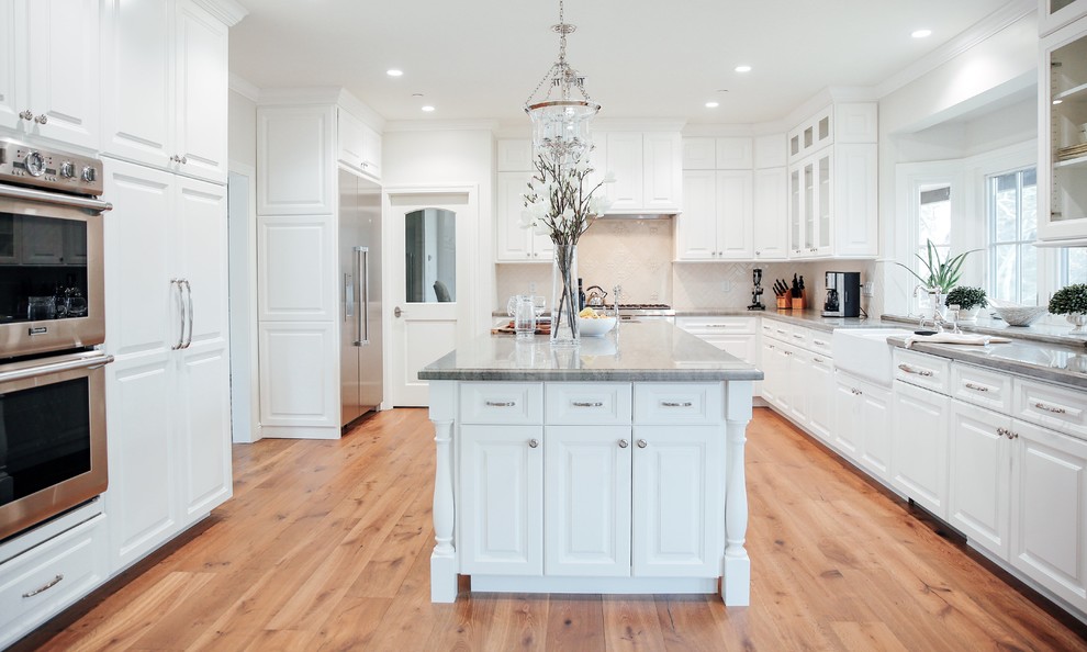 This is an example of a traditional u-shaped kitchen in Los Angeles with a farmhouse sink, raised-panel cabinets, white cabinets, beige splashback, stainless steel appliances, medium hardwood floors, with island, brown floor and grey benchtop.