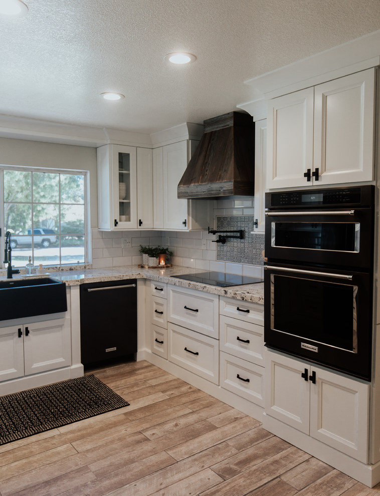 This is an example of a mid-sized country u-shaped eat-in kitchen in Los Angeles with a farmhouse sink, shaker cabinets, white cabinets, granite benchtops, white splashback, subway tile splashback, black appliances, ceramic floors, brown floor and beige benchtop.