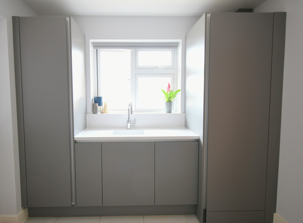 Small modern single-wall utility room in Essex with a drop-in sink, flat-panel cabinets, grey cabinets, quartzite benchtops, white walls, ceramic floors, a concealed washer and dryer, white floor and white benchtop.