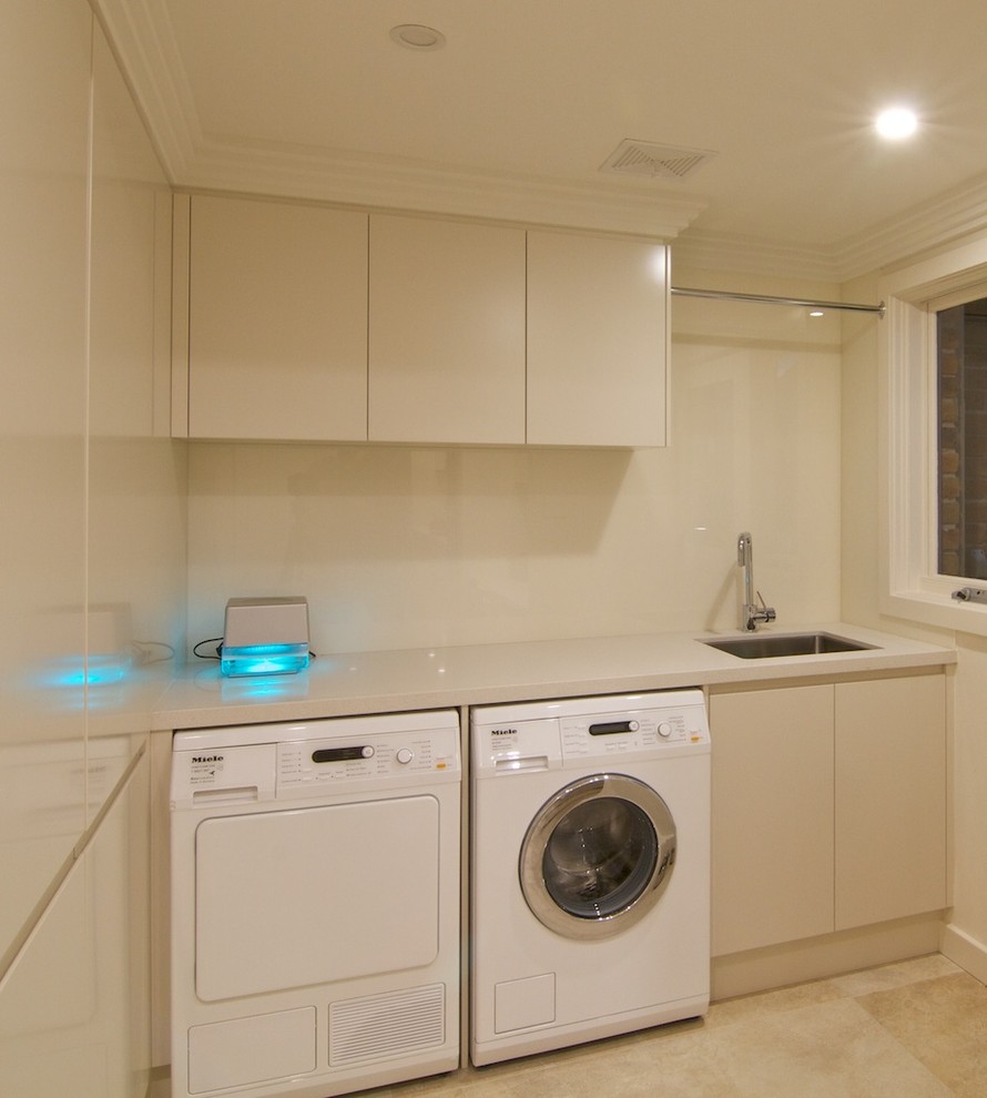 This is an example of a mid-sized contemporary galley laundry room in Sydney with an undermount sink, flat-panel cabinets, quartz benchtops, beige cabinets, brown splashback, glass sheet splashback, travertine floors, beige floor and beige benchtop.