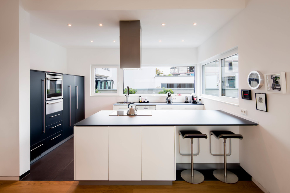 This is an example of a large contemporary galley open plan kitchen in Dusseldorf with a drop-in sink, flat-panel cabinets, white cabinets, solid surface benchtops, white splashback, glass sheet splashback, stainless steel appliances, medium hardwood floors and a peninsula.