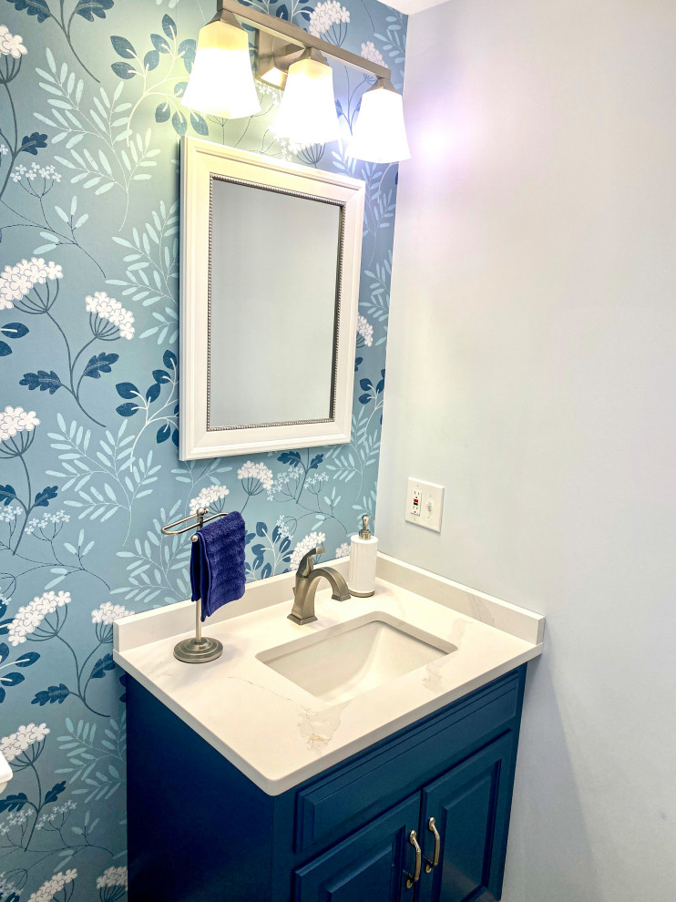 Small classic bathroom in Boston with raised-panel cabinets, blue cabinets, a two-piece toilet, blue walls, porcelain flooring, a submerged sink, engineered stone worktops, grey floors, white worktops, a single sink, a built in vanity unit and wallpapered walls.