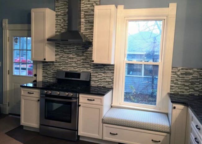 Mid-sized transitional l-shaped medium tone wood floor and brown floor kitchen pantry photo with a farmhouse sink, recessed-panel cabinets, white cabinets, granite countertops, multicolored backsplash, glass tile backsplash, stainless steel appliances, no island and black countertops