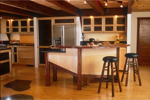 Wabi-sabi kitchen in Canada — mixed maple and walnut cabinets, glass-front uppers, handcrafted angled peninsula island, exposed timber beams, stainless fridge, wood floors