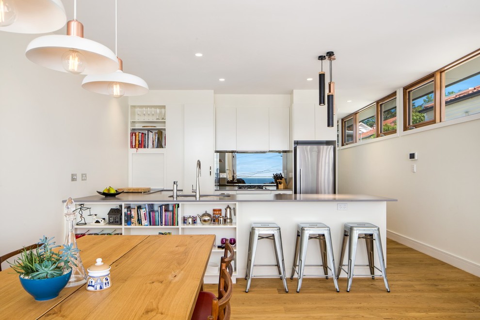 This is an example of a mid-sized contemporary galley eat-in kitchen in Sydney with light hardwood floors, an undermount sink, flat-panel cabinets, white cabinets, metallic splashback, mirror splashback and a peninsula.