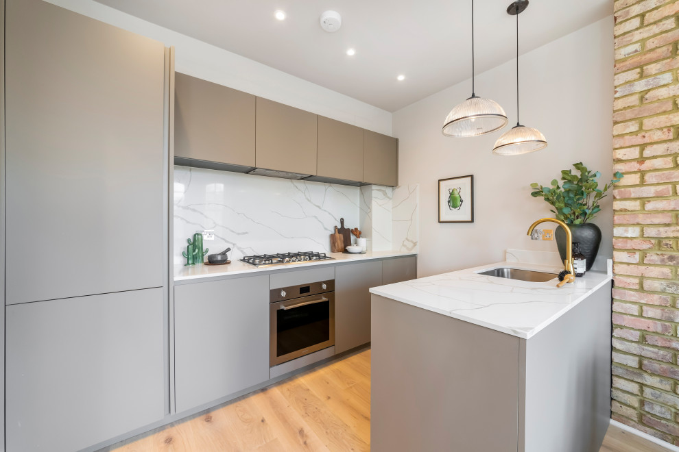 Contemporary galley kitchen in London with an undermount sink, flat-panel cabinets, grey cabinets, white splashback, stone slab splashback, medium hardwood floors, a peninsula, brown floor and white benchtop.