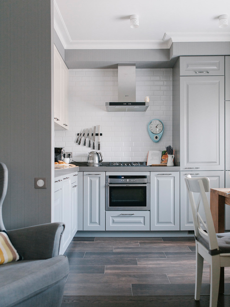 This is an example of a contemporary l-shaped open plan kitchen in Moscow with raised-panel cabinets, grey cabinets, white splashback, subway tile splashback, stainless steel appliances, dark hardwood floors, no island and brown floor.