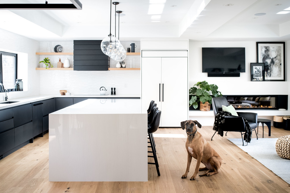 This is an example of a modern l-shaped open plan kitchen in Toronto with flat-panel cabinets, white splashback, light hardwood floors, with island, beige floor, white benchtop, an undermount sink, black cabinets and panelled appliances.