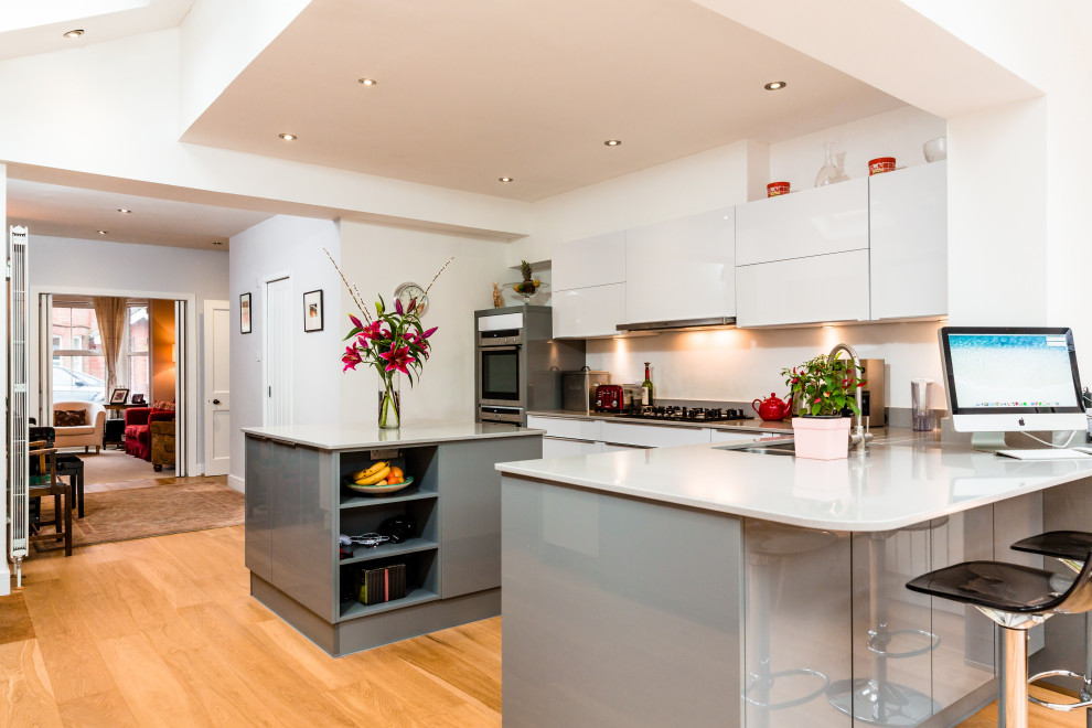 This is an example of a large contemporary l-shaped kitchen/diner in Hampshire with a double-bowl sink, flat-panel cabinets, white cabinets, quartz worktops, integrated appliances, light hardwood flooring and an island.