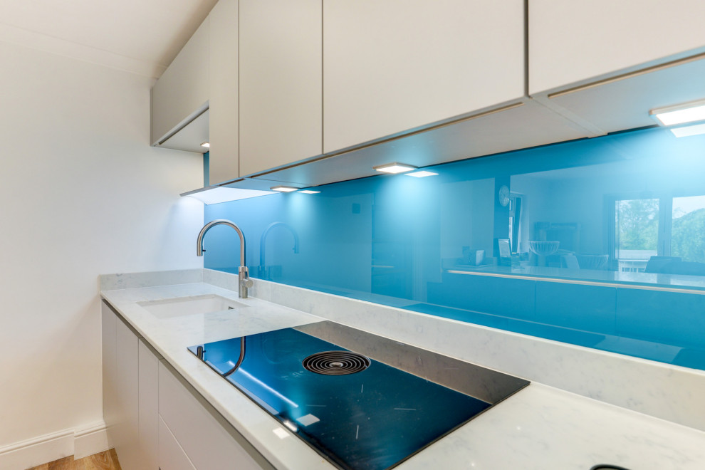 Medium sized modern grey and white single-wall kitchen/diner in Sussex with a submerged sink, flat-panel cabinets, grey cabinets, quartz worktops, blue splashback, glass sheet splashback, integrated appliances, vinyl flooring, an island, brown floors, white worktops and feature lighting.