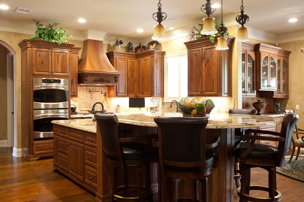 Example of a mid-sized mountain style l-shaped dark wood floor and brown floor eat-in kitchen design in Austin with raised-panel cabinets, dark wood cabinets, granite countertops, beige backsplash, ceramic backsplash, stainless steel appliances, an island, an undermount sink and beige countertops