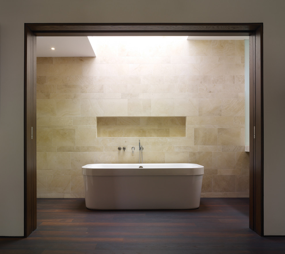 This is an example of a mid-sized modern bathroom in Los Angeles with a freestanding tub, an integrated sink, flat-panel cabinets, dark wood cabinets, a one-piece toilet, beige tile, white walls, dark hardwood floors and limestone.
