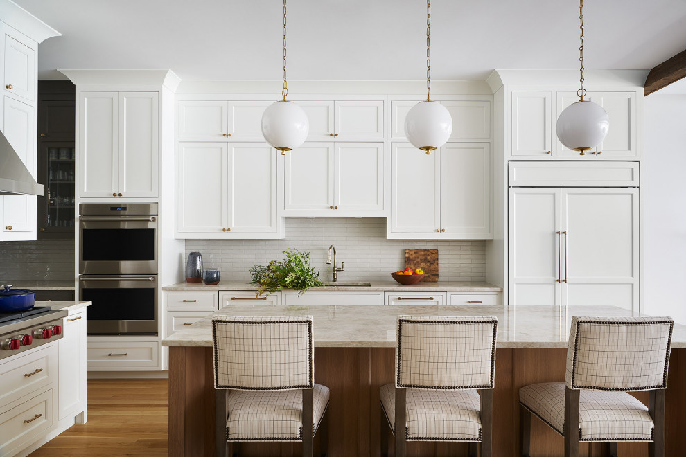 Klassisk inredning av ett beige beige l-kök, med en undermonterad diskho, skåp i shakerstil, vita skåp, vitt stänkskydd, integrerade vitvaror, mellanmörkt trägolv, en köksö och brunt golv