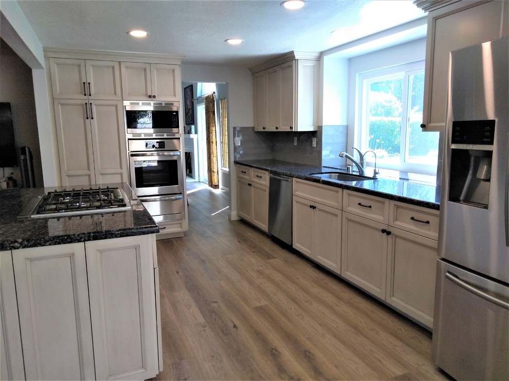 Photo of a mid-sized transitional galley open plan kitchen in Los Angeles with an undermount sink, recessed-panel cabinets, beige cabinets, granite benchtops, grey splashback, ceramic splashback, stainless steel appliances, vinyl floors, with island, brown floor and black benchtop.