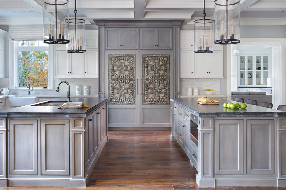 Inspiration for a transitional kitchen in New York with a farmhouse sink, recessed-panel cabinets, grey cabinets, white splashback, panelled appliances, medium hardwood floors, multiple islands, brown floor and grey benchtop.