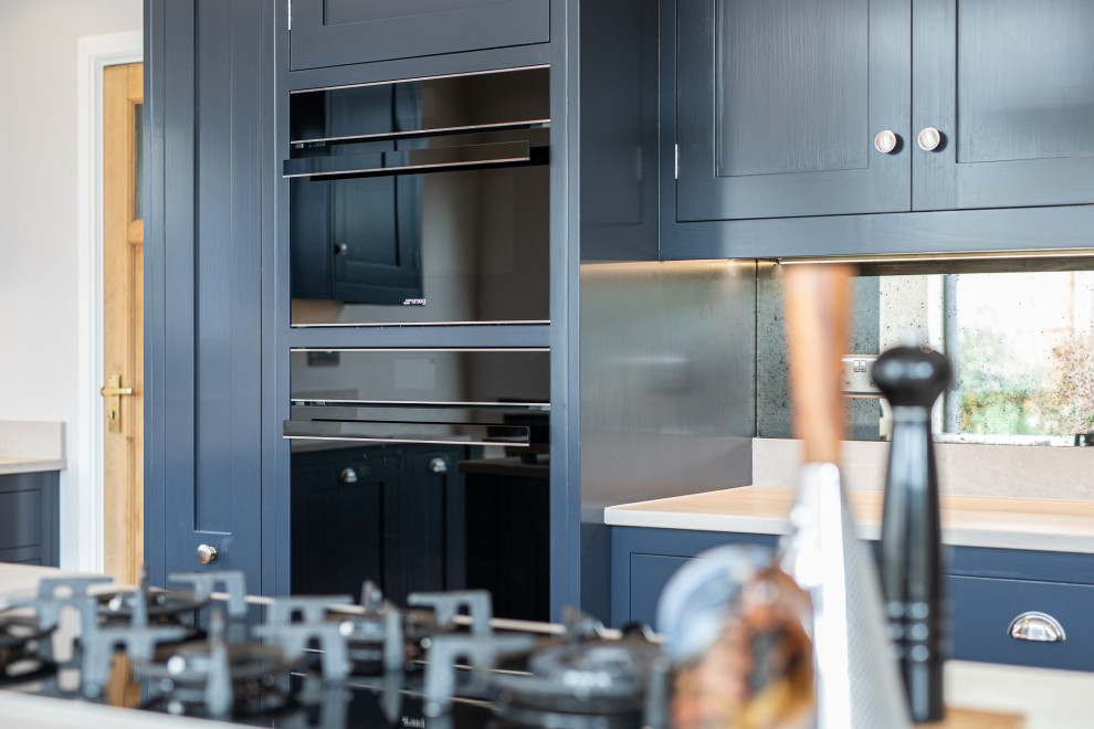 Design ideas for a large contemporary l-shaped eat-in kitchen in Hertfordshire with shaker cabinets, blue cabinets, concrete benchtops, mirror splashback, stainless steel appliances, medium hardwood floors, with island and grey benchtop.