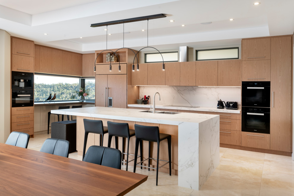 Large contemporary l-shaped open plan kitchen in Perth with a double-bowl sink, recessed-panel cabinets, light wood cabinets, white splashback, black appliances, travertine floors, with island, beige floor and white benchtop.
