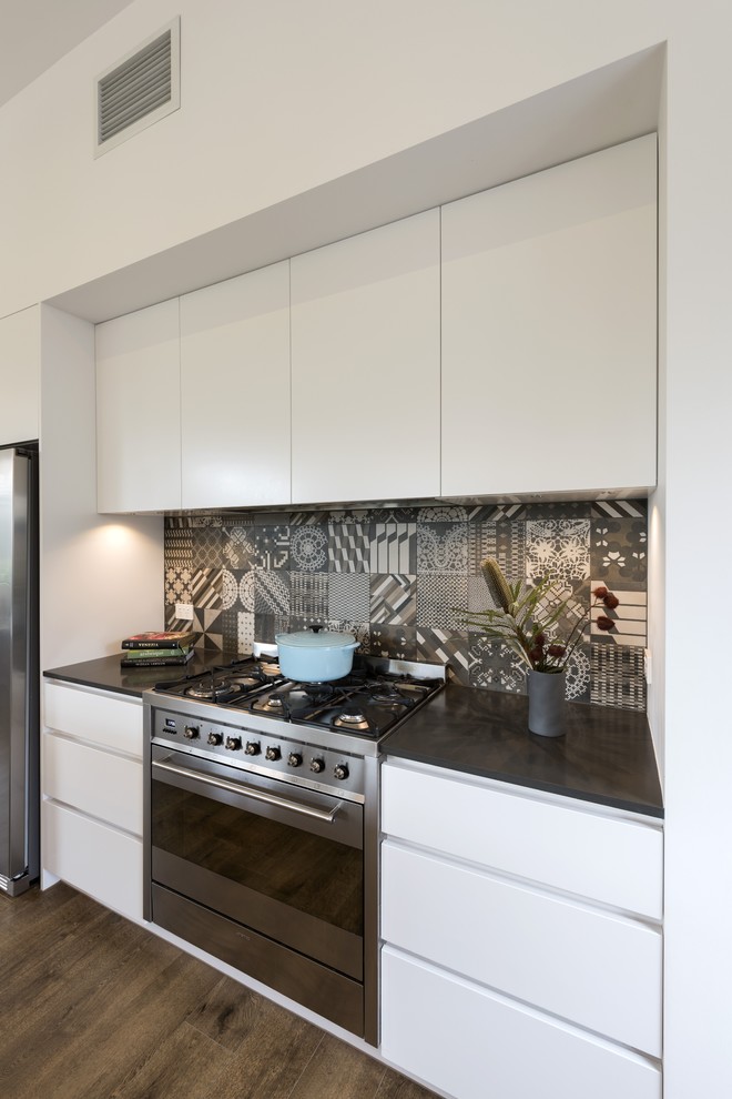 Contemporary galley open plan kitchen in Melbourne with an undermount sink, flat-panel cabinets, white cabinets, quartz benchtops, multi-coloured splashback, porcelain splashback, stainless steel appliances, porcelain floors, with island, brown floor and black benchtop.