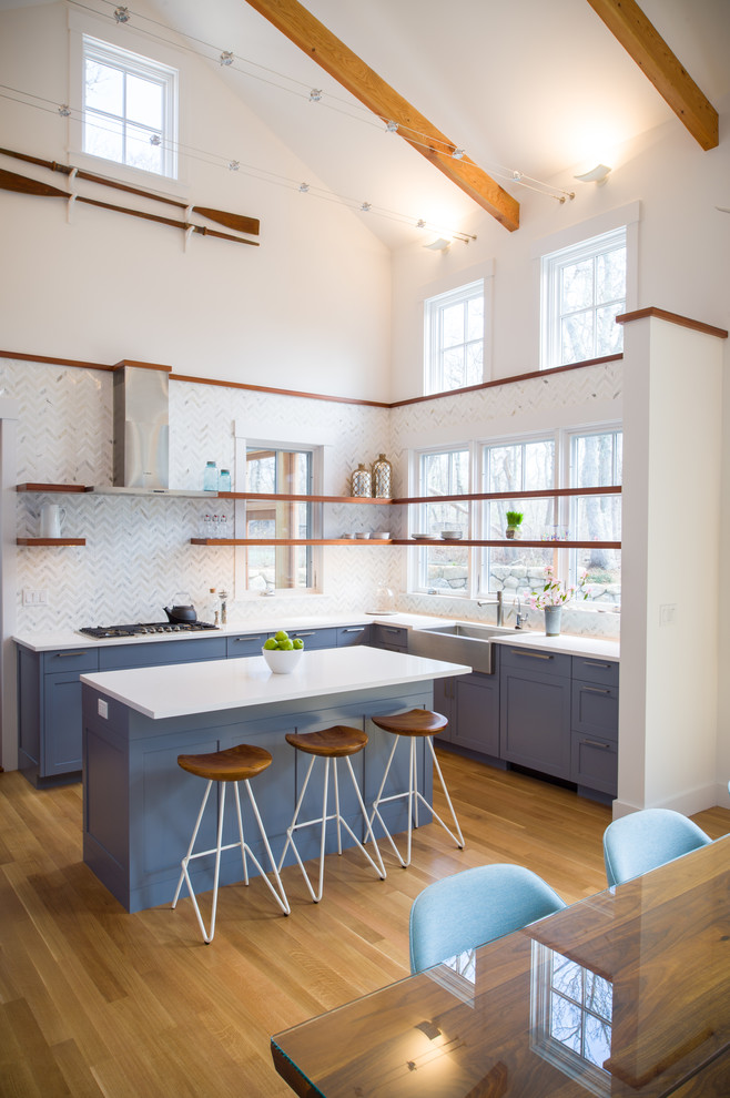 This is an example of a beach style l-shaped open plan kitchen in Boston with a farmhouse sink, shaker cabinets, blue cabinets, white splashback, marble splashback, medium hardwood floors, with island, brown floor and stainless steel appliances.