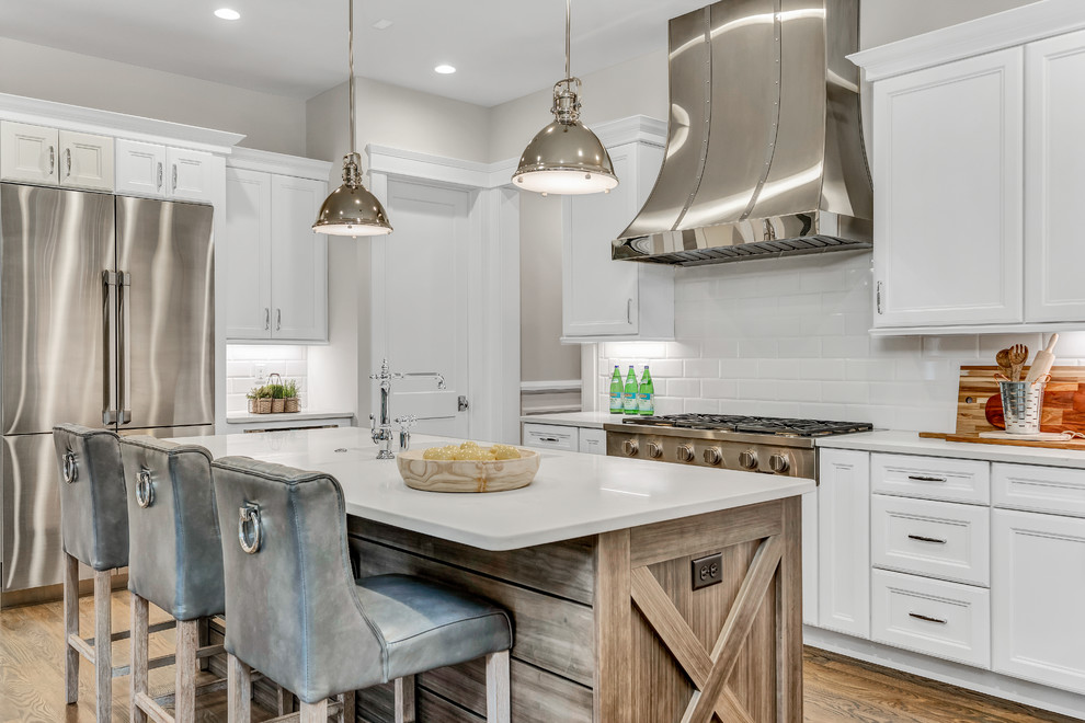 This is an example of a transitional l-shaped kitchen in St Louis with recessed-panel cabinets, white cabinets, white splashback, subway tile splashback, stainless steel appliances, dark hardwood floors, with island, brown floor and white benchtop.