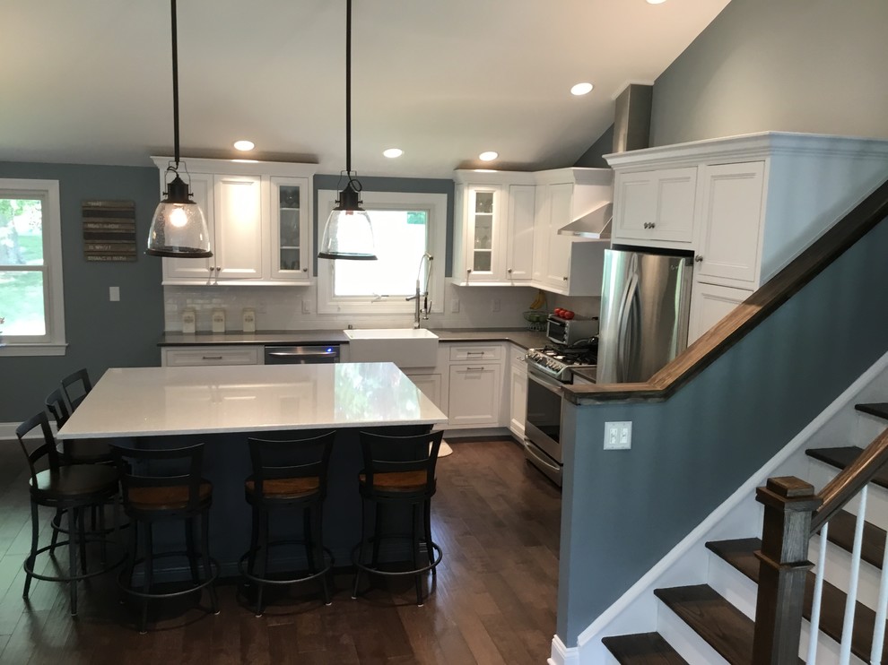 Example of a mid-sized transitional l-shaped dark wood floor and brown floor kitchen design in New York with a farmhouse sink, recessed-panel cabinets, white cabinets, granite countertops, white backsplash, subway tile backsplash, stainless steel appliances, an island and black countertops
