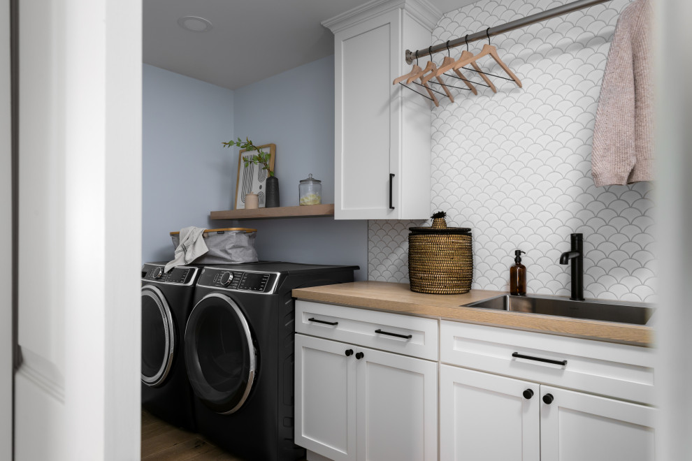 Foto på en stor skandinavisk bruna linjär tvättstuga enbart för tvätt, med en nedsänkt diskho, skåp i shakerstil, vita skåp, träbänkskiva, vitt stänkskydd, stänkskydd i keramik, blå väggar, ljust trägolv och en tvättmaskin och torktumlare bredvid varandra