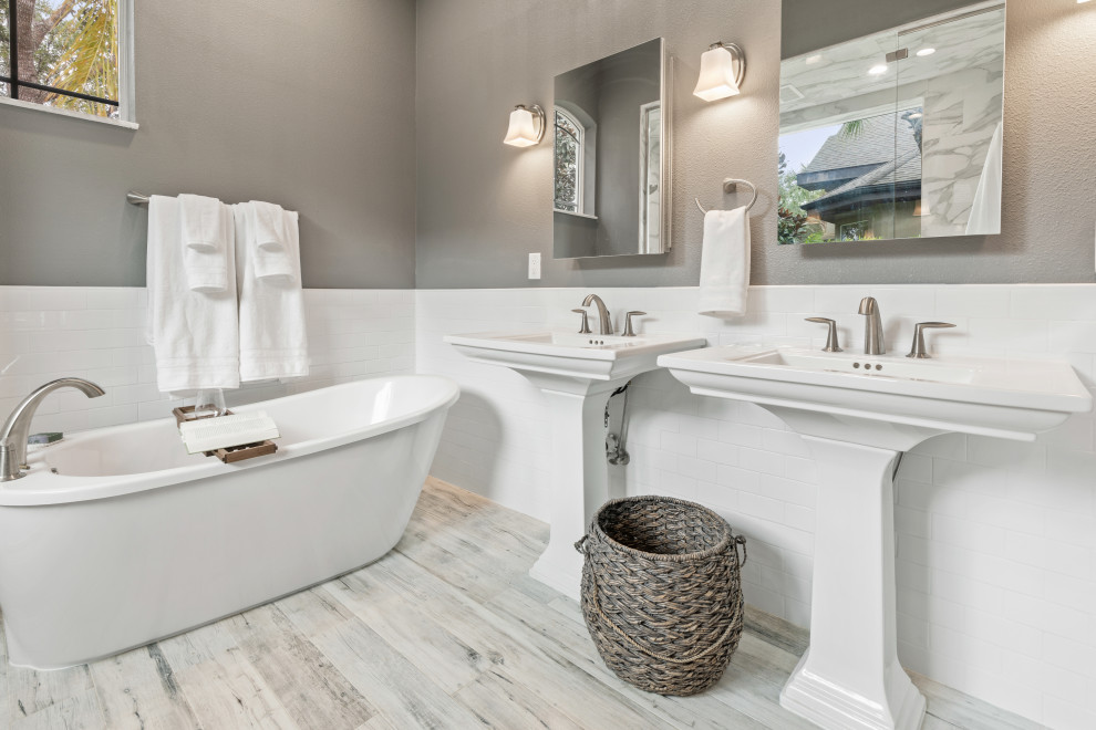 This is an example of a transitional master bathroom in Orlando with a freestanding tub, a one-piece toilet, white tile, grey walls, wood-look tile, a pedestal sink, grey floor, a shower seat, a double vanity and a freestanding vanity.