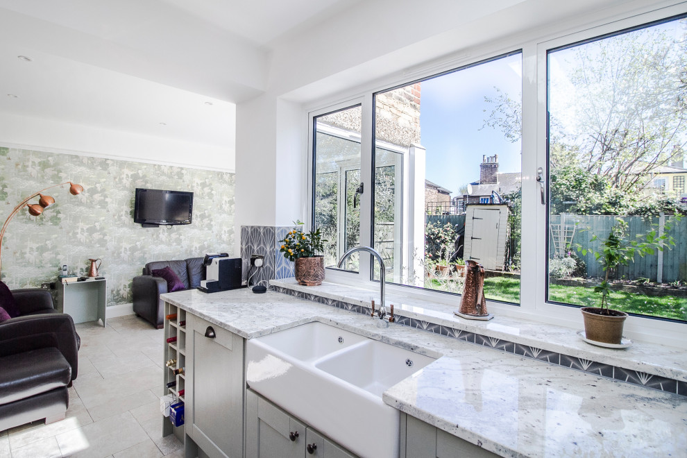 Photo of a mid-sized transitional l-shaped open plan kitchen in London with a farmhouse sink, shaker cabinets, granite benchtops, blue splashback, ceramic splashback, stainless steel appliances, limestone floors, with island, grey floor and white benchtop.
