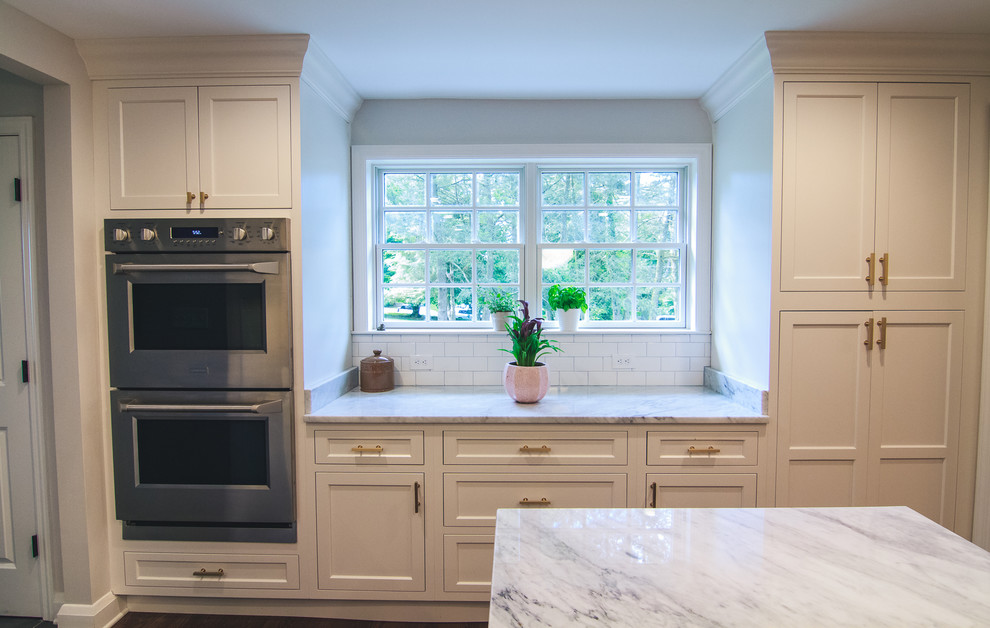 Photo of a large traditional u-shaped open plan kitchen in Baltimore with a farmhouse sink, shaker cabinets, white cabinets, marble benchtops, white splashback, subway tile splashback, stainless steel appliances, medium hardwood floors, with island and brown floor.