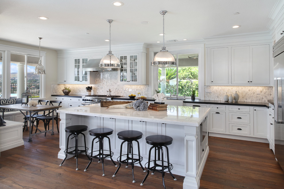 This is an example of a large beach style l-shaped eat-in kitchen in Orange County with a farmhouse sink, beaded inset cabinets, white cabinets, marble benchtops, white splashback, mosaic tile splashback, stainless steel appliances, medium hardwood floors and with island.
