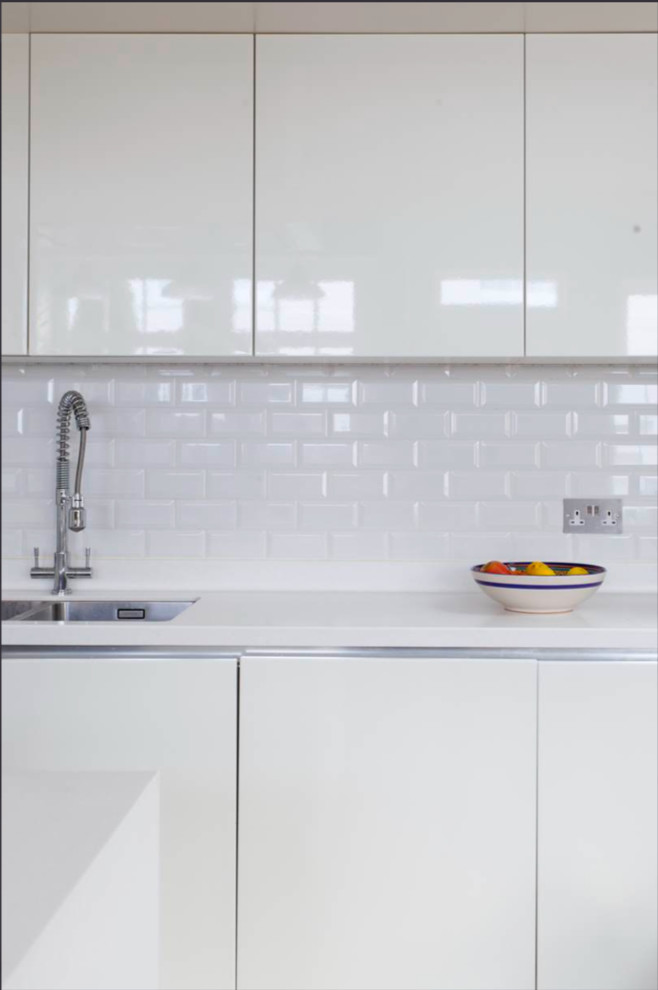 This is an example of a small contemporary galley open plan kitchen in London with white cabinets, solid surface benchtops, white splashback and with island.
