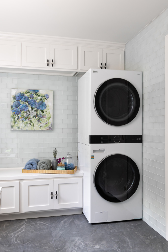 Photo of a transitional galley dedicated laundry room in Kansas City with recessed-panel cabinets, white cabinets, quartz benchtops, blue splashback, glass tile splashback, porcelain floors, a stacked washer and dryer, grey floor and white benchtop.