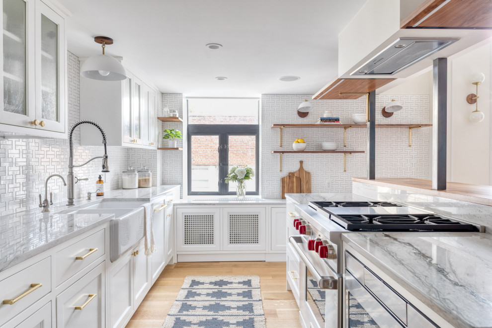 Mid-sized transitional galley eat-in kitchen in New York with a farmhouse sink, glass-front cabinets, white cabinets, quartzite benchtops, white splashback, ceramic splashback, stainless steel appliances, light hardwood floors, with island, brown floor and grey benchtop.