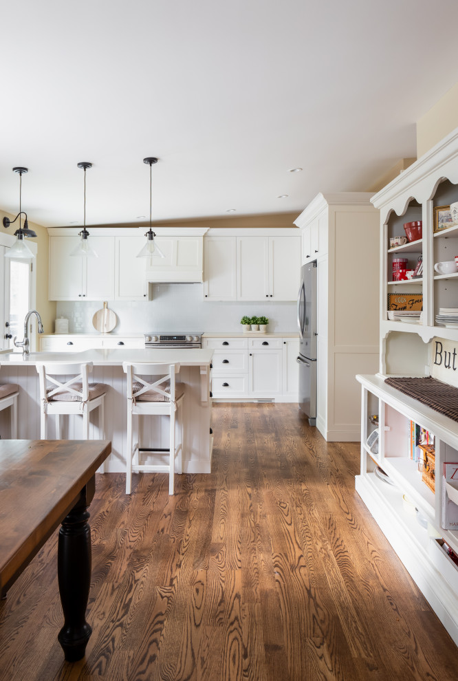 Example of a large mountain style l-shaped medium tone wood floor and brown floor eat-in kitchen design in Vancouver with a farmhouse sink, shaker cabinets, white cabinets, quartzite countertops, white backsplash, subway tile backsplash, stainless steel appliances, an island and white countertops