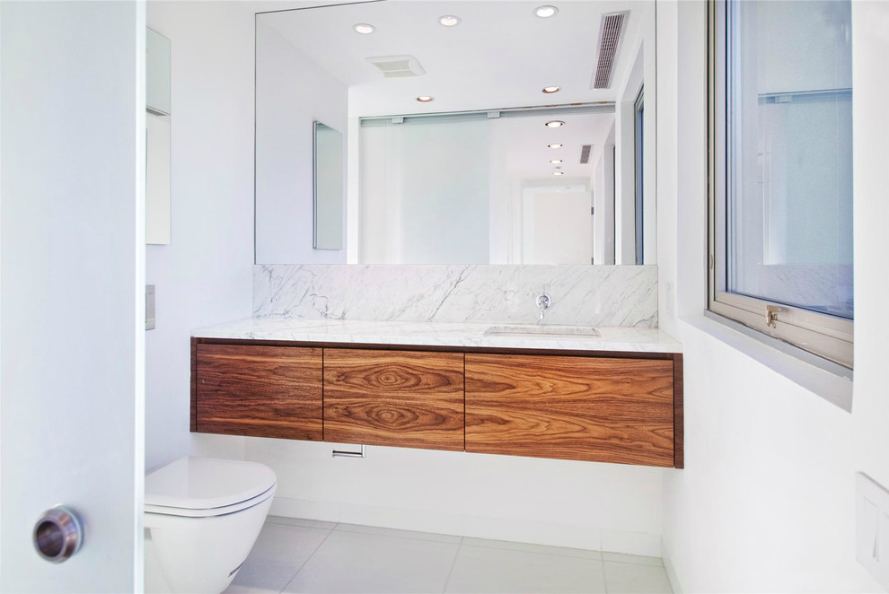 Inspiration for a small contemporary powder room in New York with flat-panel cabinets, medium wood cabinets, a wall-mount toilet, white tile, stone slab, white walls, ceramic floors, an undermount sink, marble benchtops and white floor.
