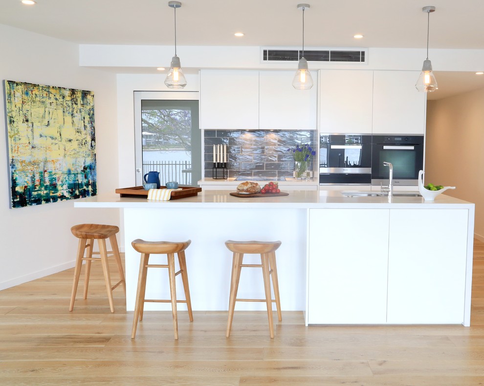 Design ideas for a contemporary galley kitchen in Brisbane with flat-panel cabinets, white cabinets, grey splashback, light hardwood floors and with island.