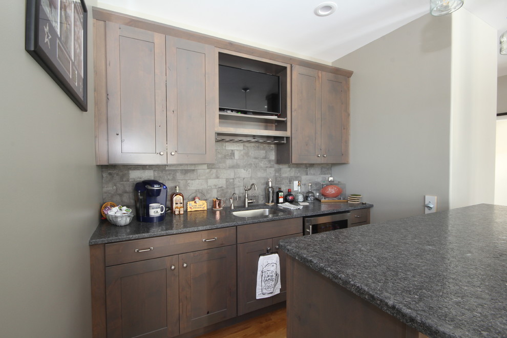 This is an example of a mid-sized country galley seated home bar in Manchester with an undermount sink, shaker cabinets, dark wood cabinets, granite benchtops, grey splashback, stone tile splashback, medium hardwood floors, blue floor and black benchtop.