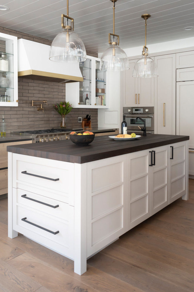Inspiration for a mid-sized country l-shaped open plan kitchen in Chicago with an undermount sink, glass-front cabinets, white cabinets, stainless steel benchtops, brown splashback, ceramic splashback, coloured appliances, light hardwood floors, multiple islands, beige floor and wood.