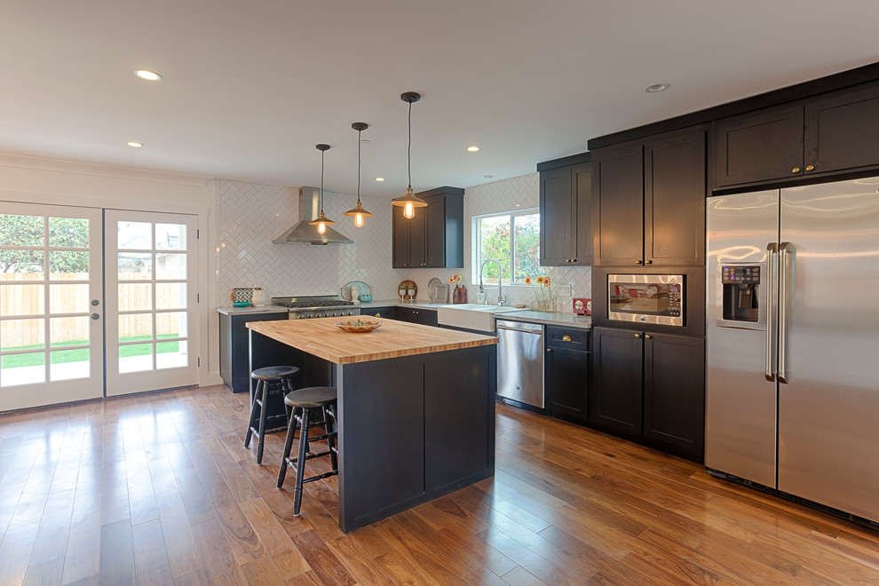 Open concept kitchen - mid-sized modern l-shaped medium tone wood floor open concept kitchen idea with a farmhouse sink, shaker cabinets, black cabinets, marble countertops, white backsplash, ceramic backsplash, stainless steel appliances and an island