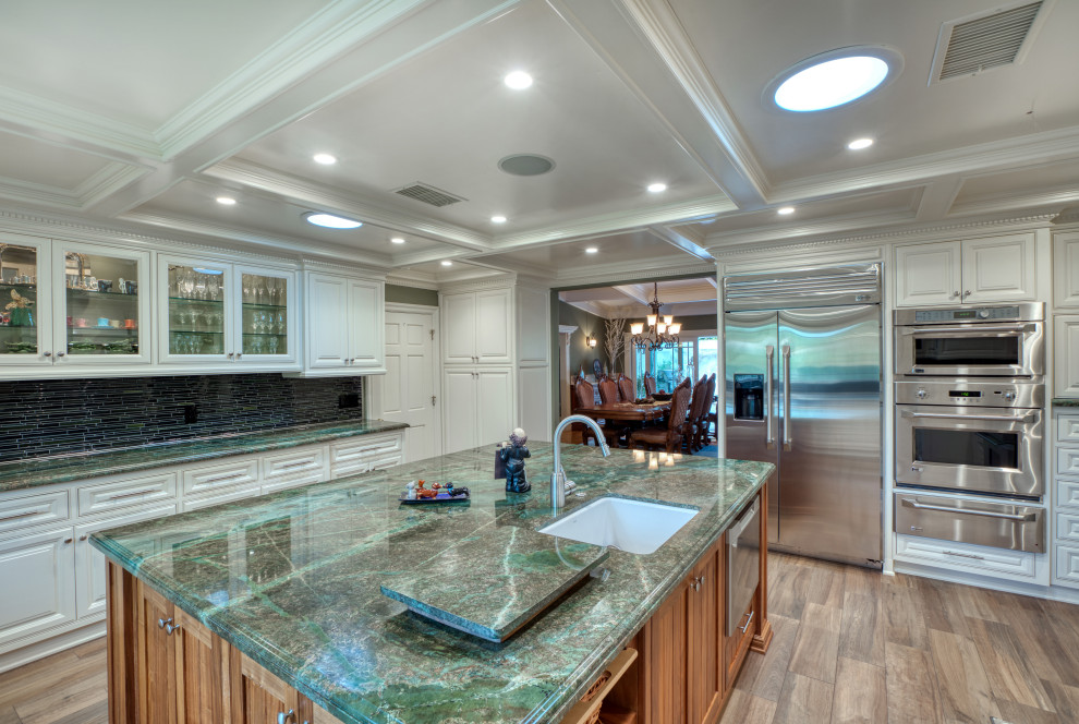 This is an example of a large traditional u-shaped eat-in kitchen in Los Angeles with a farmhouse sink, raised-panel cabinets, white cabinets, granite benchtops, green splashback, matchstick tile splashback, stainless steel appliances, light hardwood floors, with island, beige floor and green benchtop.