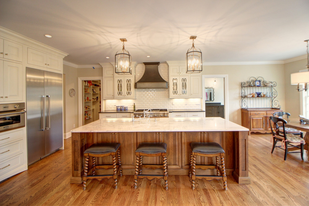 Open concept kitchen - large transitional l-shaped medium tone wood floor open concept kitchen idea in Denver with a farmhouse sink, white cabinets, granite countertops, white backsplash, subway tile backsplash, stainless steel appliances, an island and glass-front cabinets