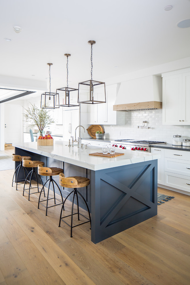 Photo of a beach style galley kitchen in Los Angeles with a farmhouse sink, shaker cabinets, white cabinets, white splashback, panelled appliances, with island, black benchtop, light hardwood floors and beige floor.