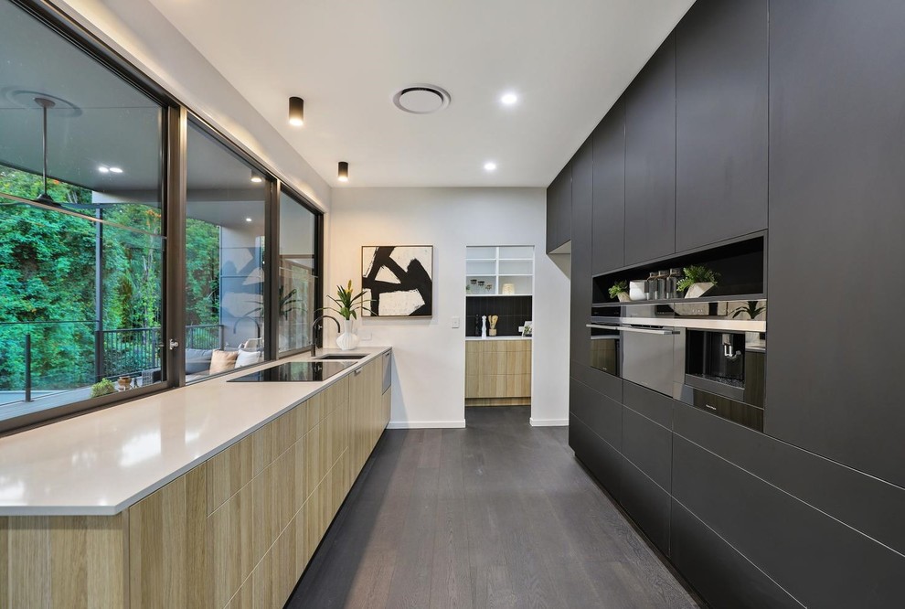 Photo of a contemporary galley kitchen in Sunshine Coast with an undermount sink, flat-panel cabinets, black cabinets, window splashback, black appliances, medium hardwood floors, brown floor and white benchtop.