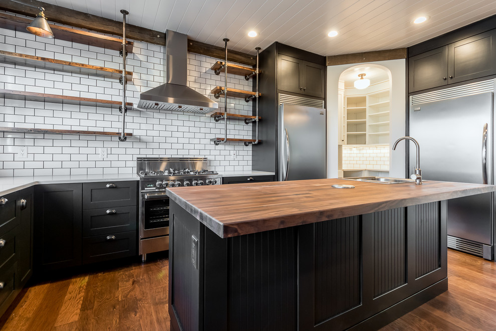 Inspiration for a large industrial u-shaped medium tone wood floor and brown floor kitchen remodel in Chicago with shaker cabinets, black cabinets, wood countertops, white backsplash, subway tile backsplash, stainless steel appliances, an island and a farmhouse sink