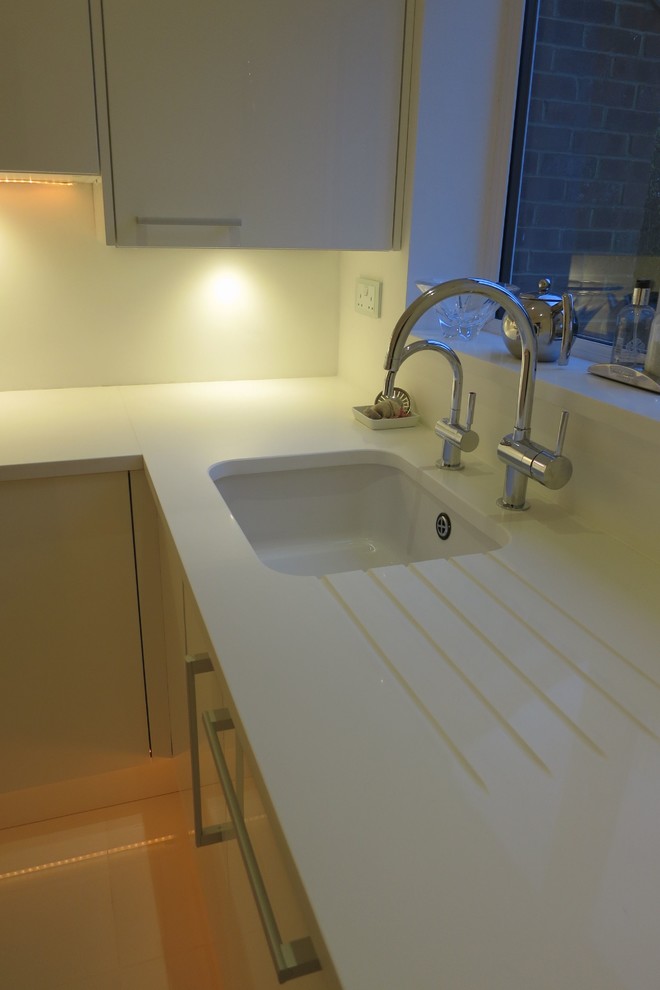 Photo of a small modern u-shaped separate kitchen in Hampshire with an undermount sink, flat-panel cabinets, white cabinets, quartzite benchtops, white splashback, glass sheet splashback, white appliances, ceramic floors and no island.
